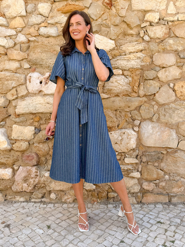 Model wears a blue pinstripe midi shirt dress with tie waist and short sleeves, paired with white strappy heels against a…