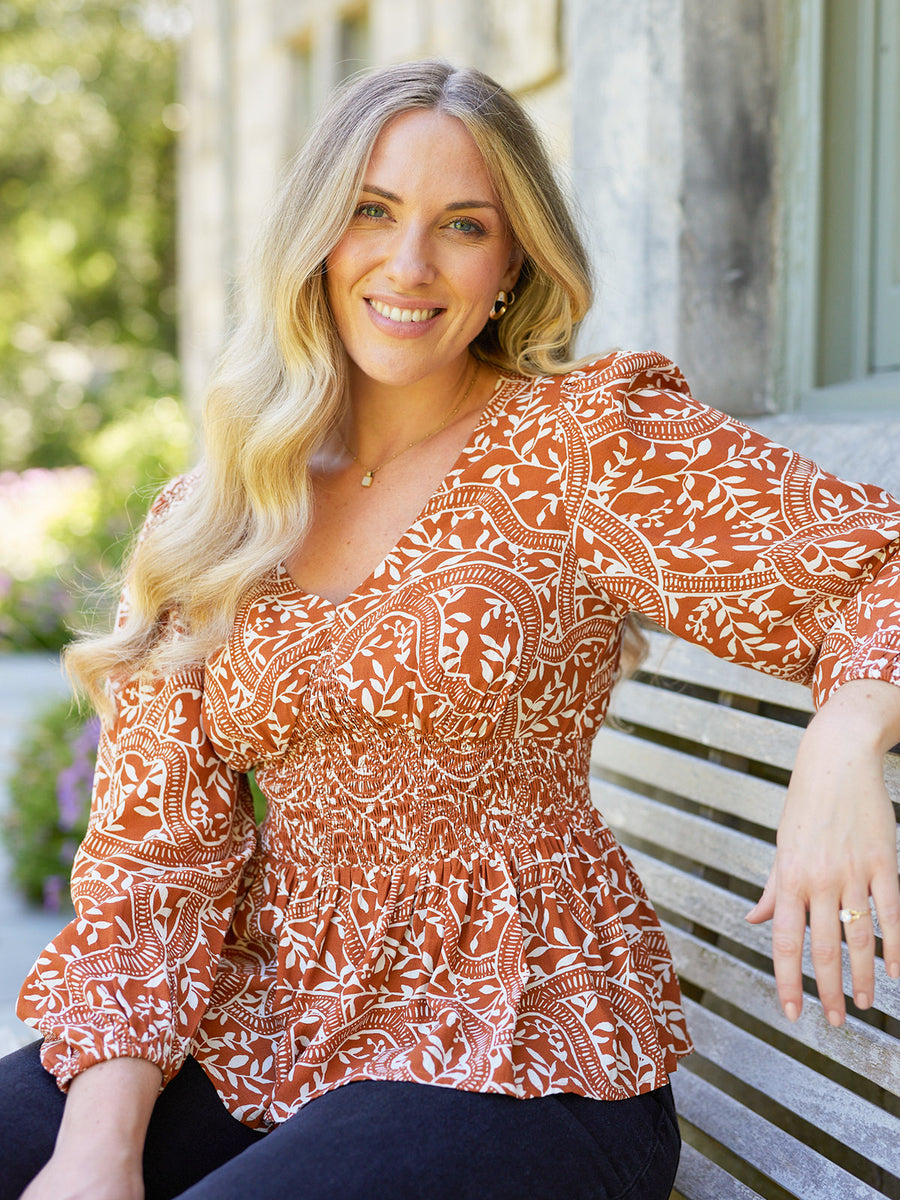 Model seated outdoors wearing a rust orange and white floral print smocked peplum top with three-quarter sleeves