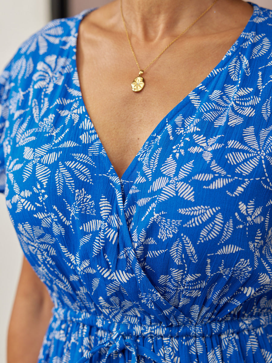 Close-up of a model wearing a blue wrap dress with a white leaf print, cap sleeves, and elasticated waist, styled with a…