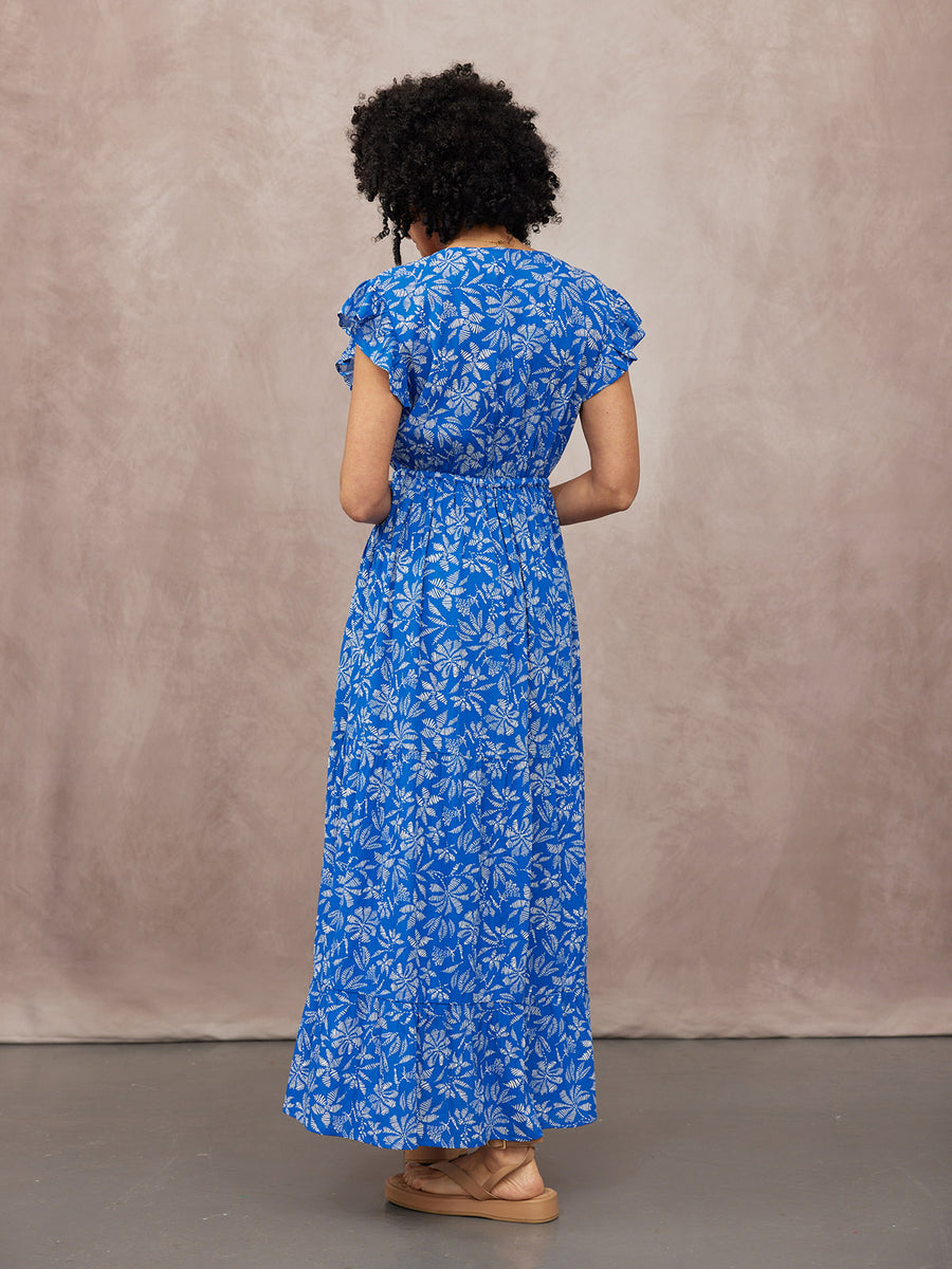 Model seen from behind in a blue maxi dress with white floral print, ruffle sleeves, and elasticated waist, paired with tan…