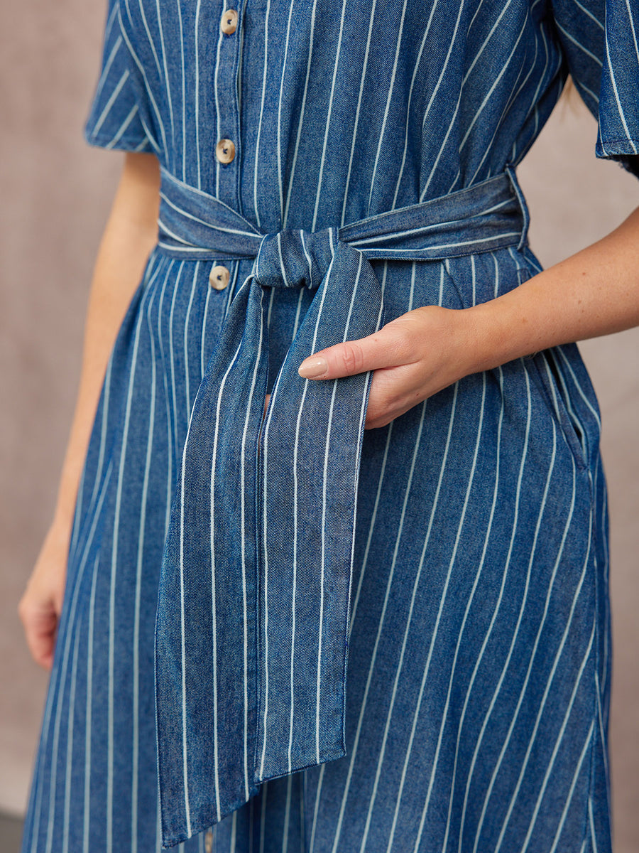 Close-up of a model wearing a blue pinstripe denim midi dress with flutter sleeves, button front, and a tied waist belt…