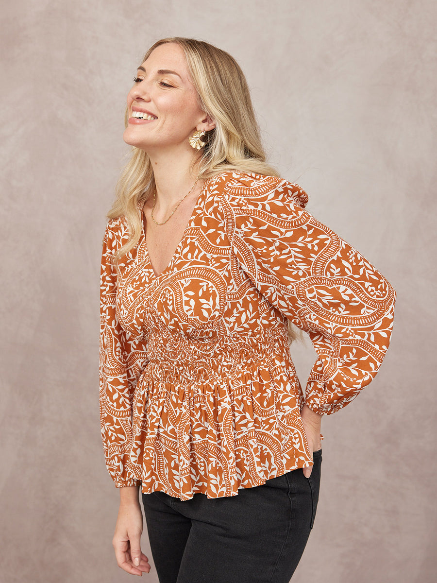 Model wears a rust orange and white floral print peplum blouse with V-neck, ruffle collar, smocked waist