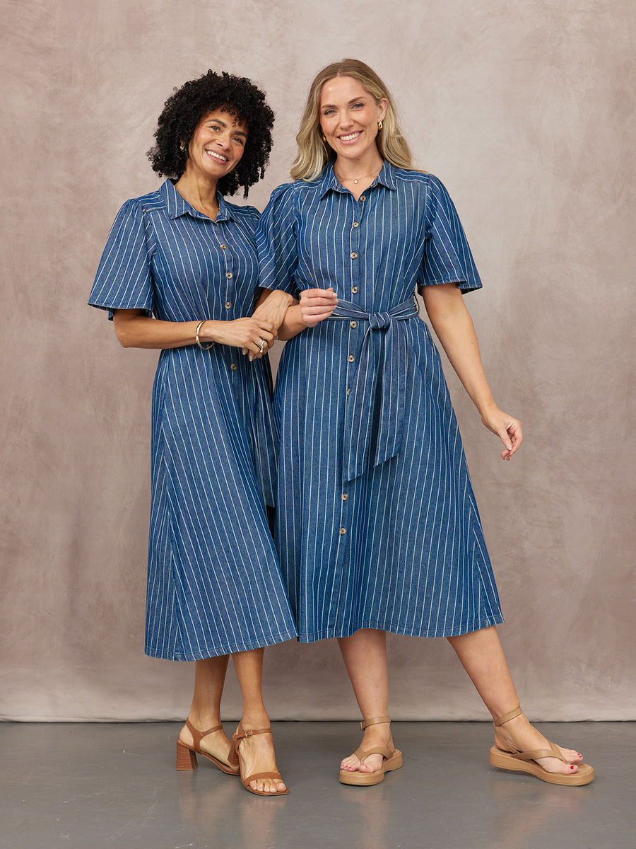 Two models wearing blue pinstripe midi shirt dresses with short sleeves and tan sandals, one belted at the waist, against a…