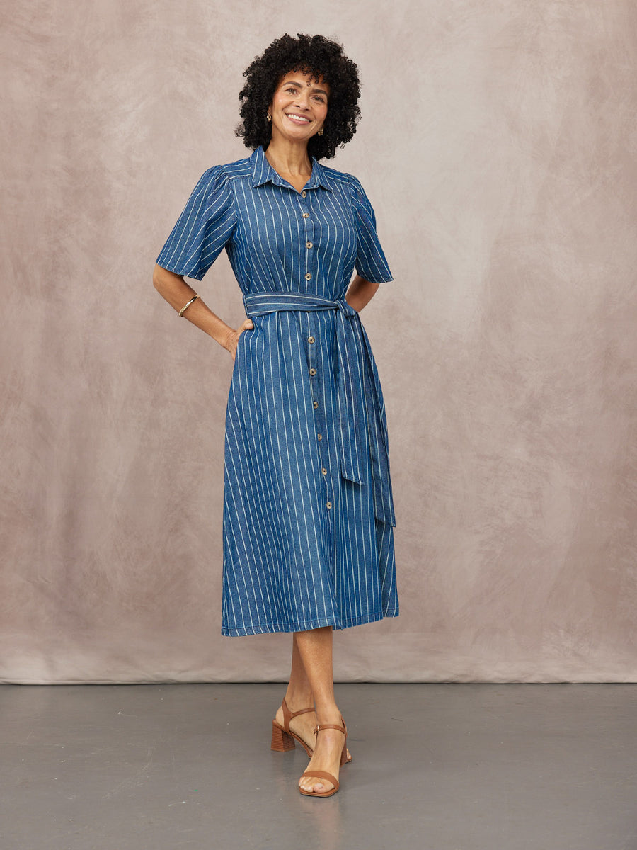 Model wearing a blue denim pinstripe midi shirtdress with short sleeves, button front, and tie belt, styled with tan…