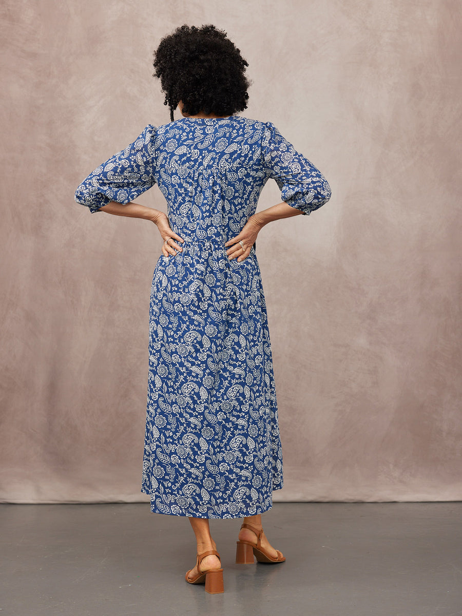 Model seen from behind wearing a blue and white paisley midi dress with puff sleeves, tan block-heel sandals, studio…