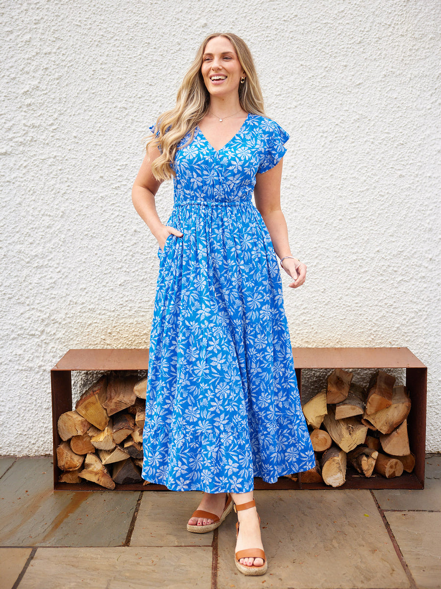 Model wearing a blue floral maxi dress with cap sleeves and V-neck, paired with tan wedge sandals, outdoors.