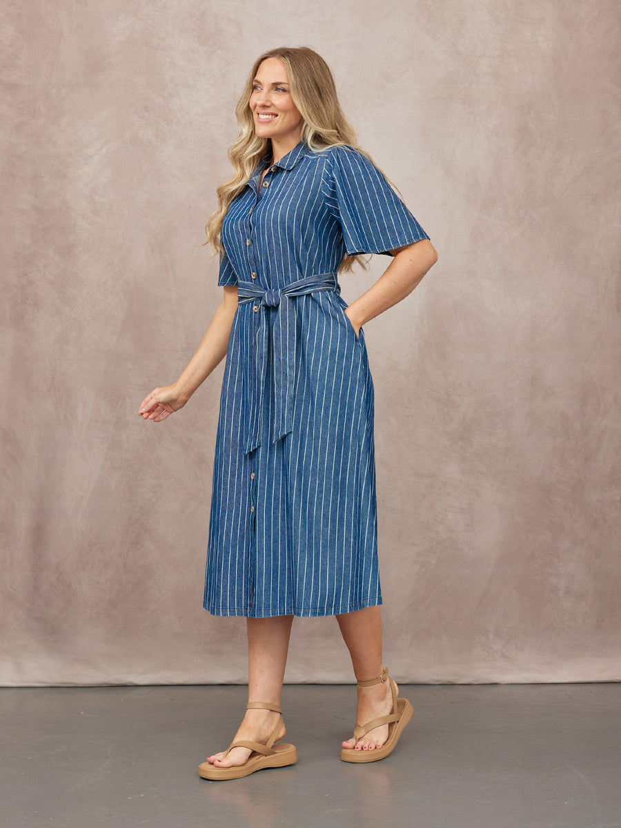 Model wearing a blue pinstripe midi shirt dress with short sleeves, tie belt, and tan flatform sandals against a dusty pink…