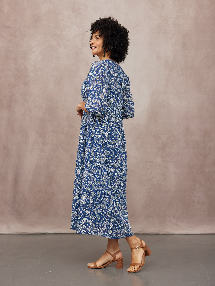 Model wears a blue paisley-print midi dress with long sleeves and a cinched waist, paired with tan block-heel sandals.