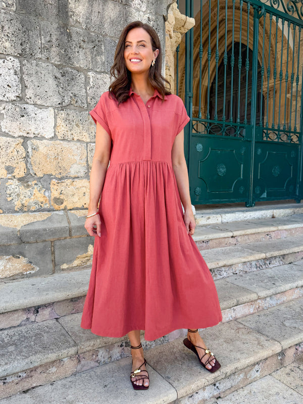 Model wearing a terracotta short-sleeve shirt midi dress with a gathered skirt, paired with dark strappy sandals on stone…