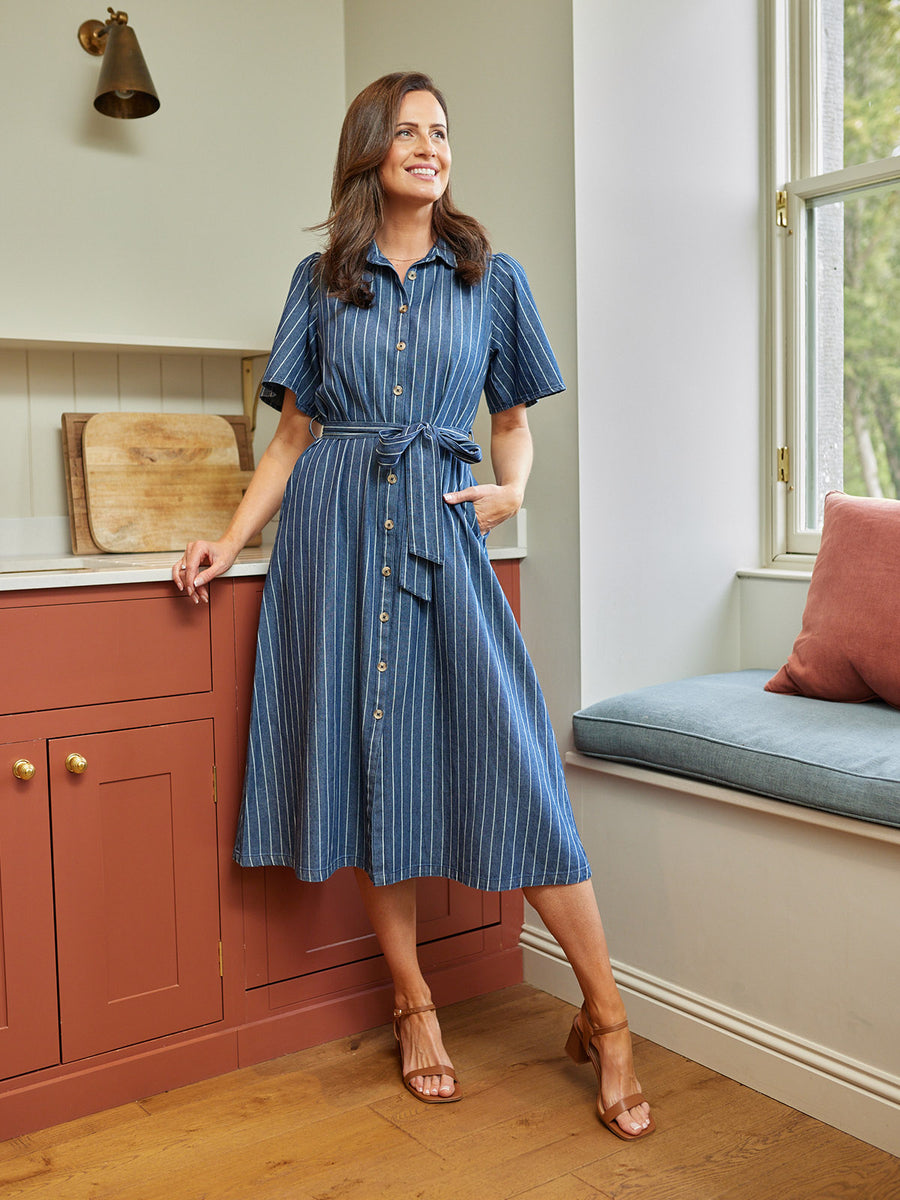 Model wearing a blue striped midi shirt dress with short sleeves, tie belt, and buttons, styled with tan heeled sandals in…