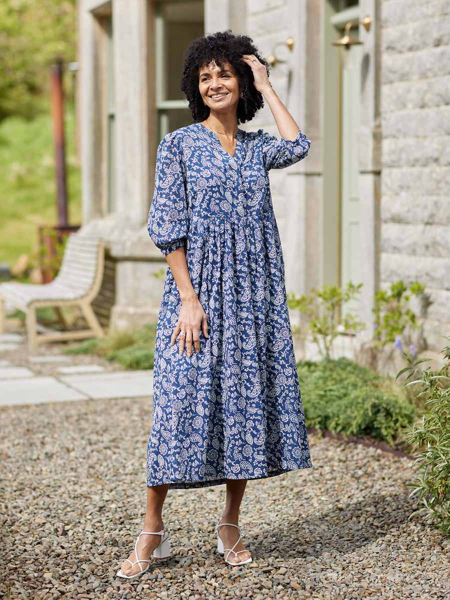 Model wearing a blue and white paisley midi dress with puff sleeves, V-neck, and gathered skirt, styled with white heeled…