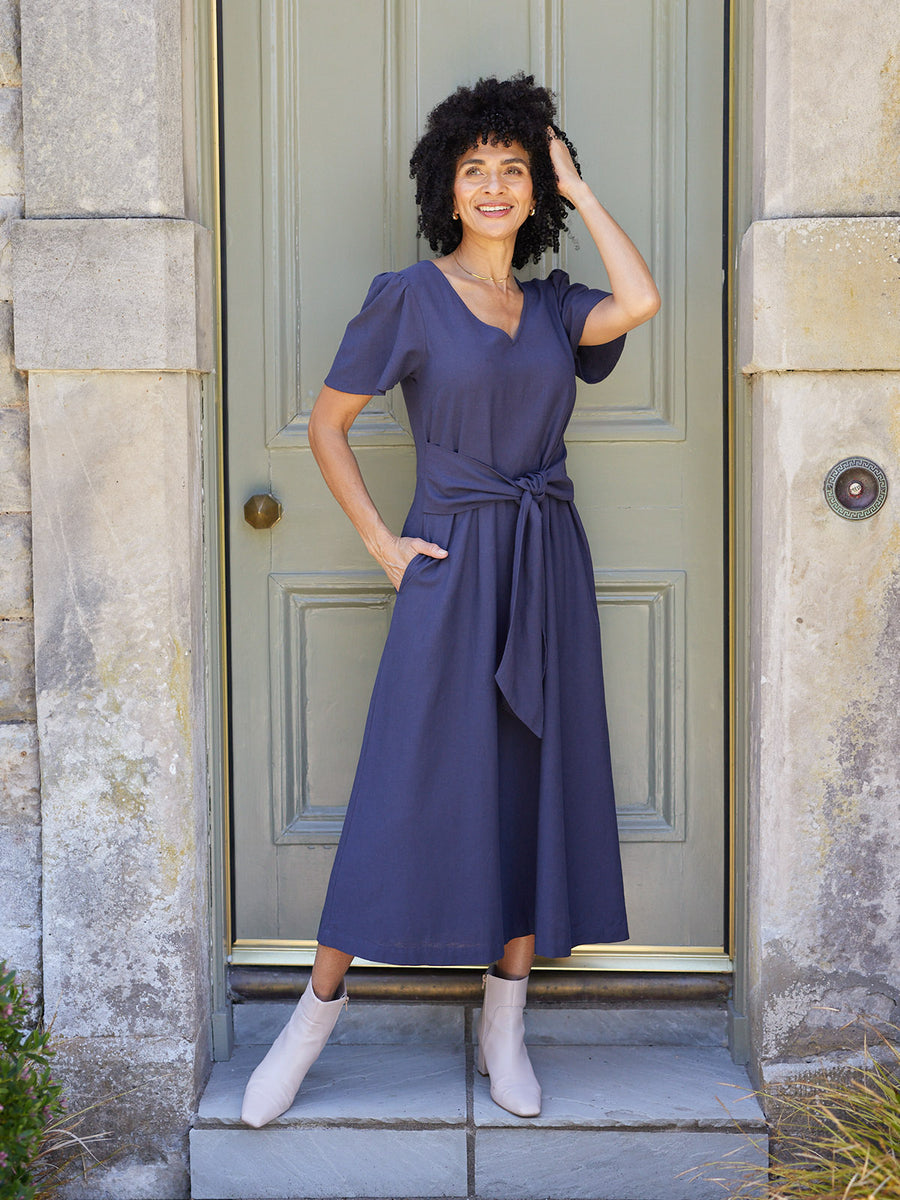Model wearing a navy midi dress with puff sleeves, V-neck, and tie waist, styled with white ankle boots outside