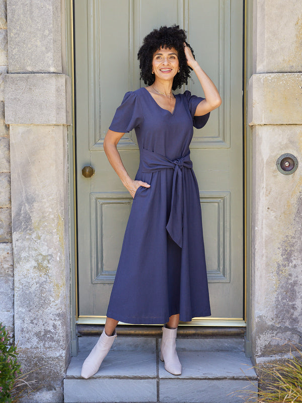 Model wearing a navy midi dress with puff sleeves, V-neck, and tie waist, styled with white ankle boots outside