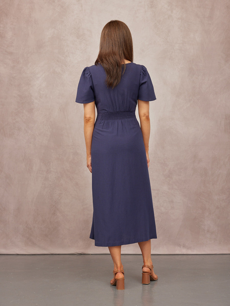 Model seen from behind wearing a navy midi dress with puff sleeves and shirred waist, paired with tan block-heel sandals.