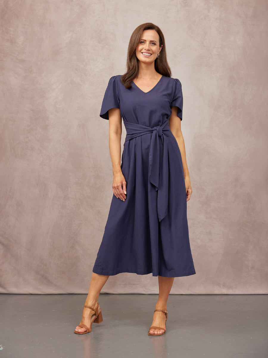 Model wearing a navy midi dress with a V-neck, flutter sleeves, and a self-tie waist belt, styled with tan block-heels