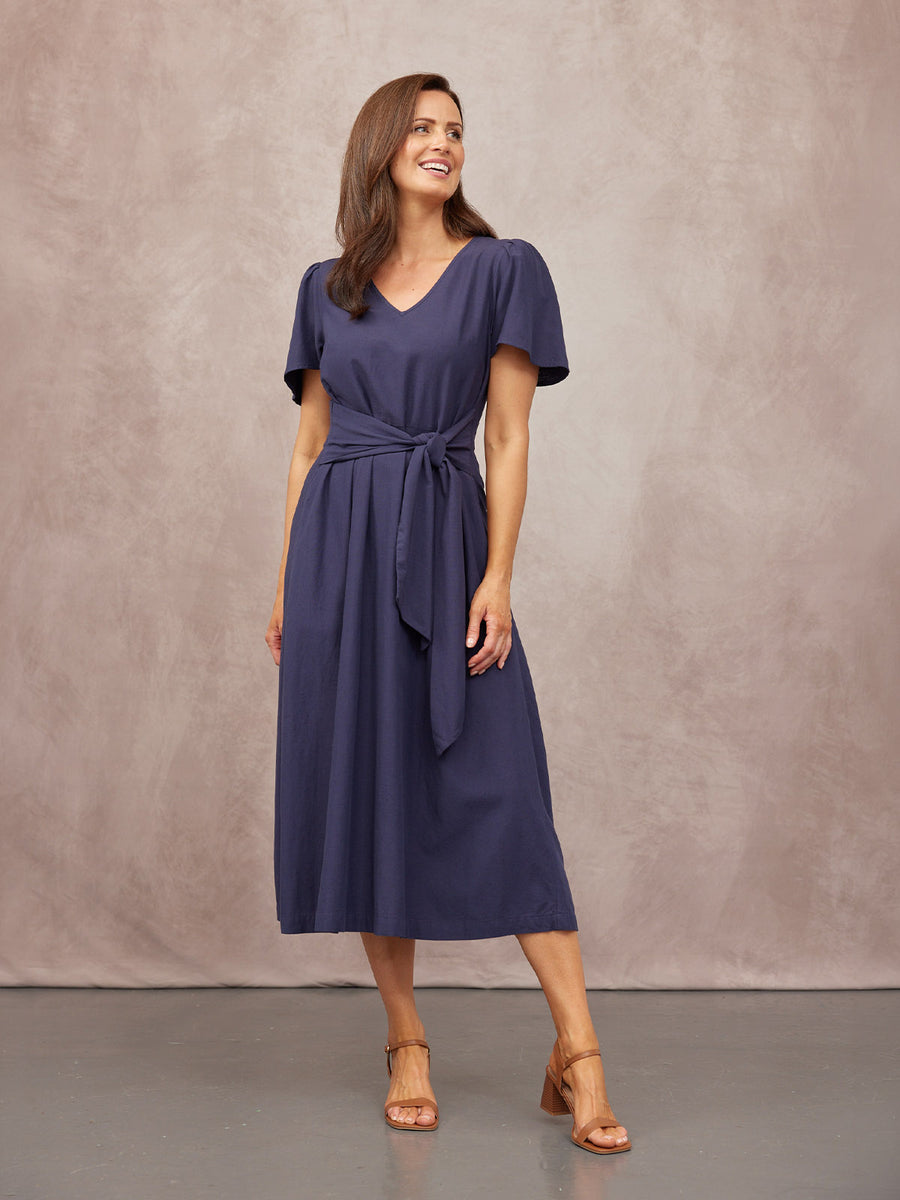 Model wearing a navy blue midi dress with V-neck, flutter sleeves, and a tie waist belt, styled with tan block-heel sandals.