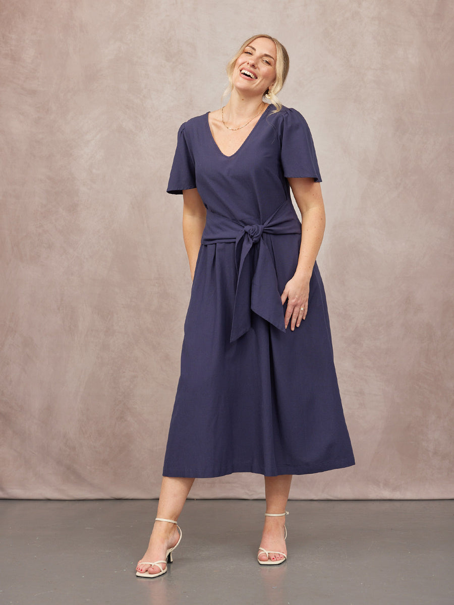 Model wearing a navy blue midi dress with a V-neckline, flutter sleeves, and a tie waist, styled with white strappy heels.