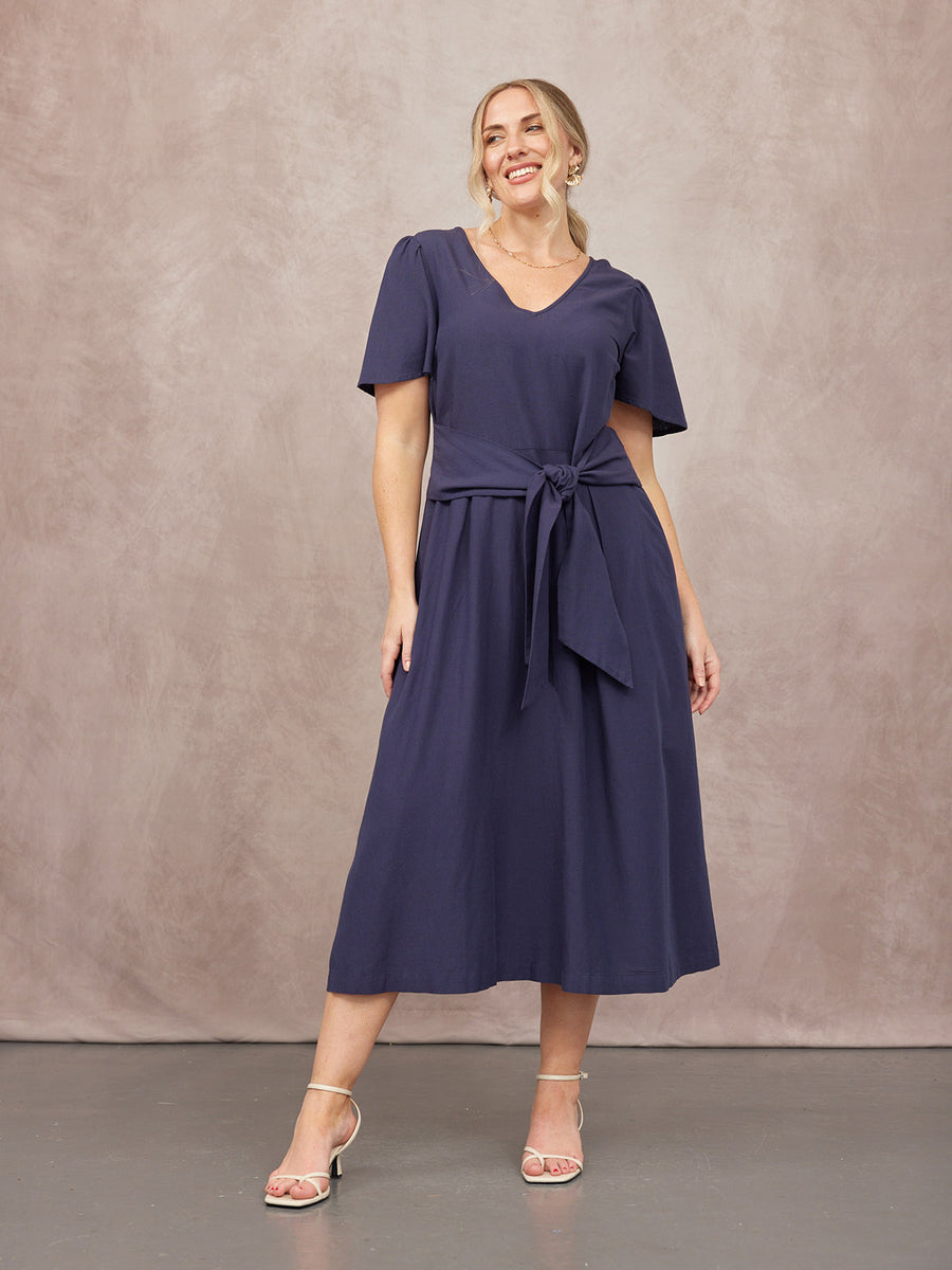 Model wearing a navy blue midi dress with flutter sleeves, V-neckline, and a self-tie waist bow, paired with white shoes