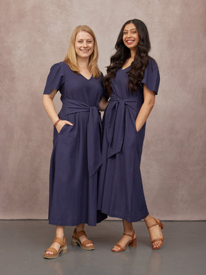 Two models wearing matching navy midi dresses with flutter sleeves, tie waist, and pockets, paired with tan sandals.