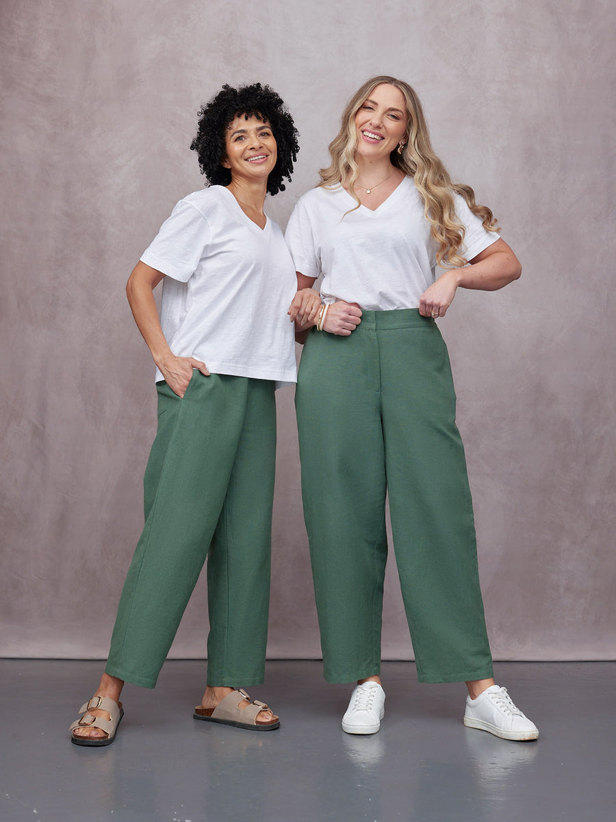 Two women wearing green Myrtle Barrel Leg Trousers styled with white v-neck tees, sandals and white trainers in a studio sett