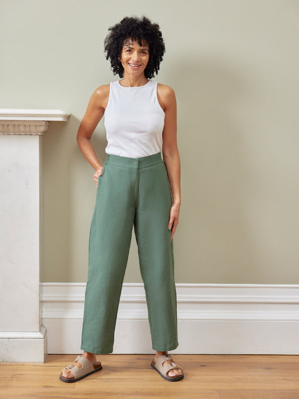 Myrtle Barrel Leg Trouser in green linen, styled with a white sleeveless top and tan sandals on a smiling woman indoors.