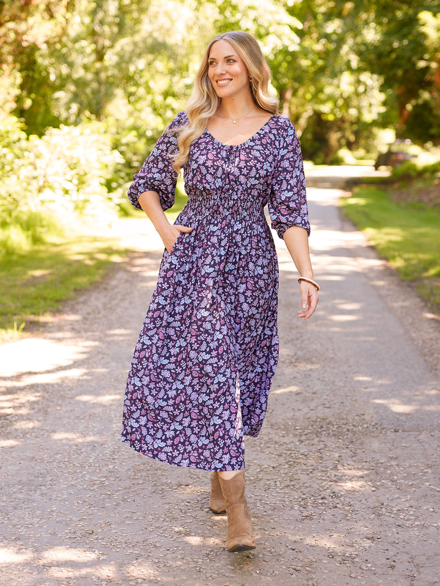 Model walks a sunlit gravel path in a navy floral midi dress with smocked waist, puff sleeves, and tan knee-high boots.