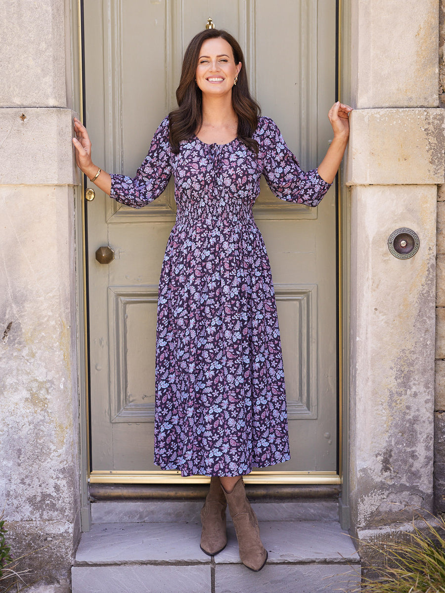 Model wearing a dark purple floral midi dress with shirred waist and three-quarter sleeves, paired with tan ankle boots.