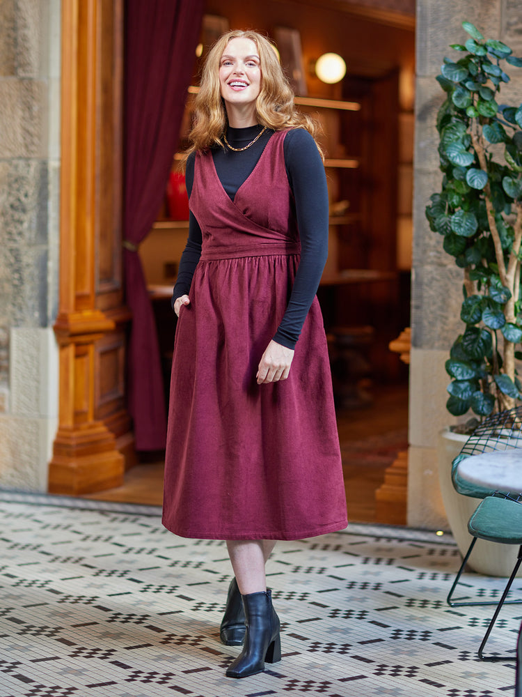 A model wears the burgundy Ally dress whilst standing in a well lit room beside a tall plant