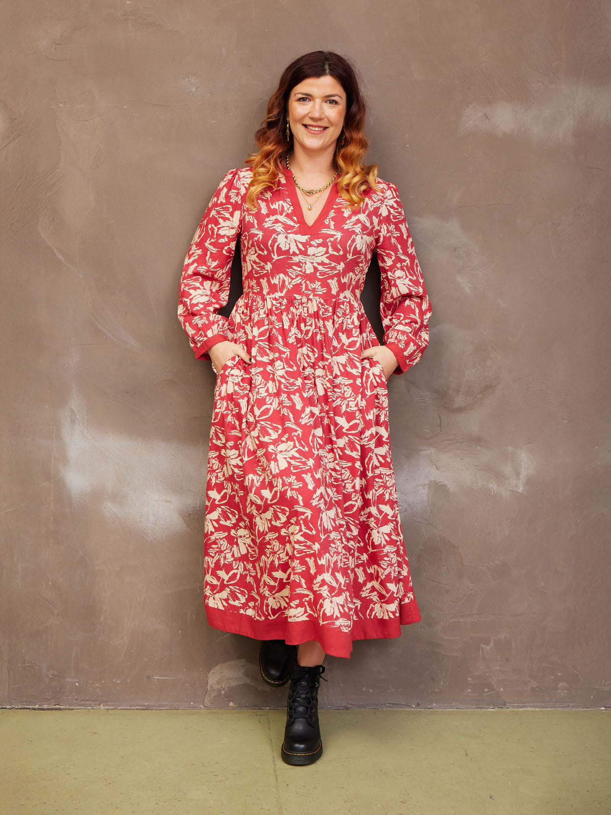Woman in a floral red dress with hands in pockets, smiling against a textured wall.