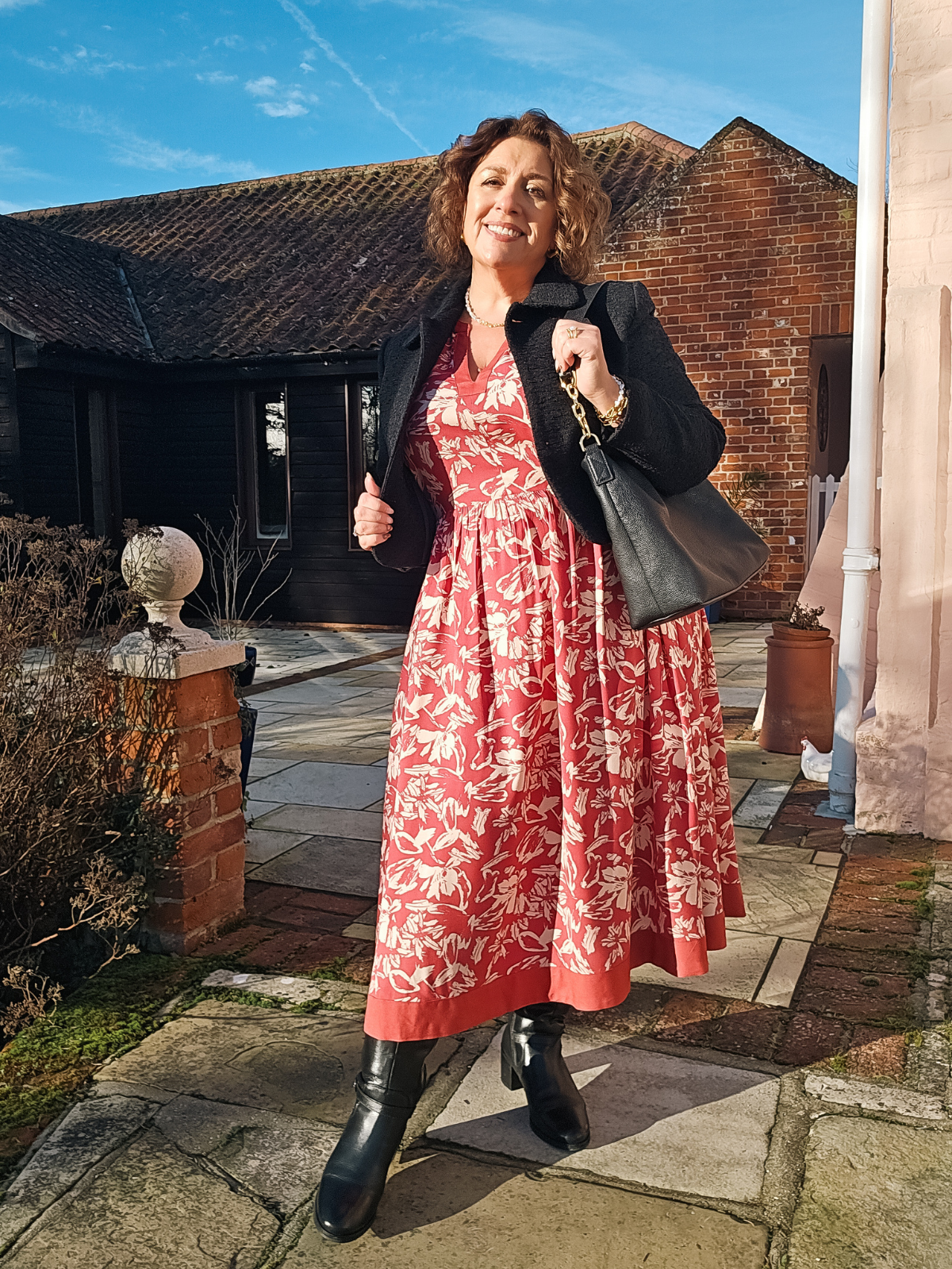 Woman in red floral dress and black boots, standing outside a rustic building.