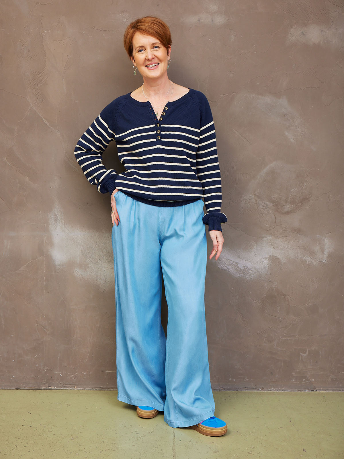 A person in a striped sweater and light blue pants standing against a textured wall.