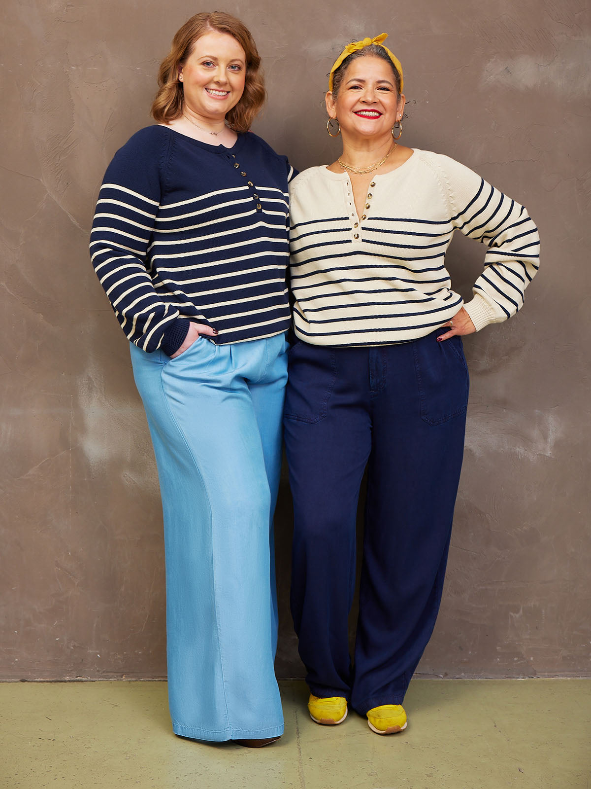 Two people wearing striped sweaters and wide-leg pants smiling against a neutral background.