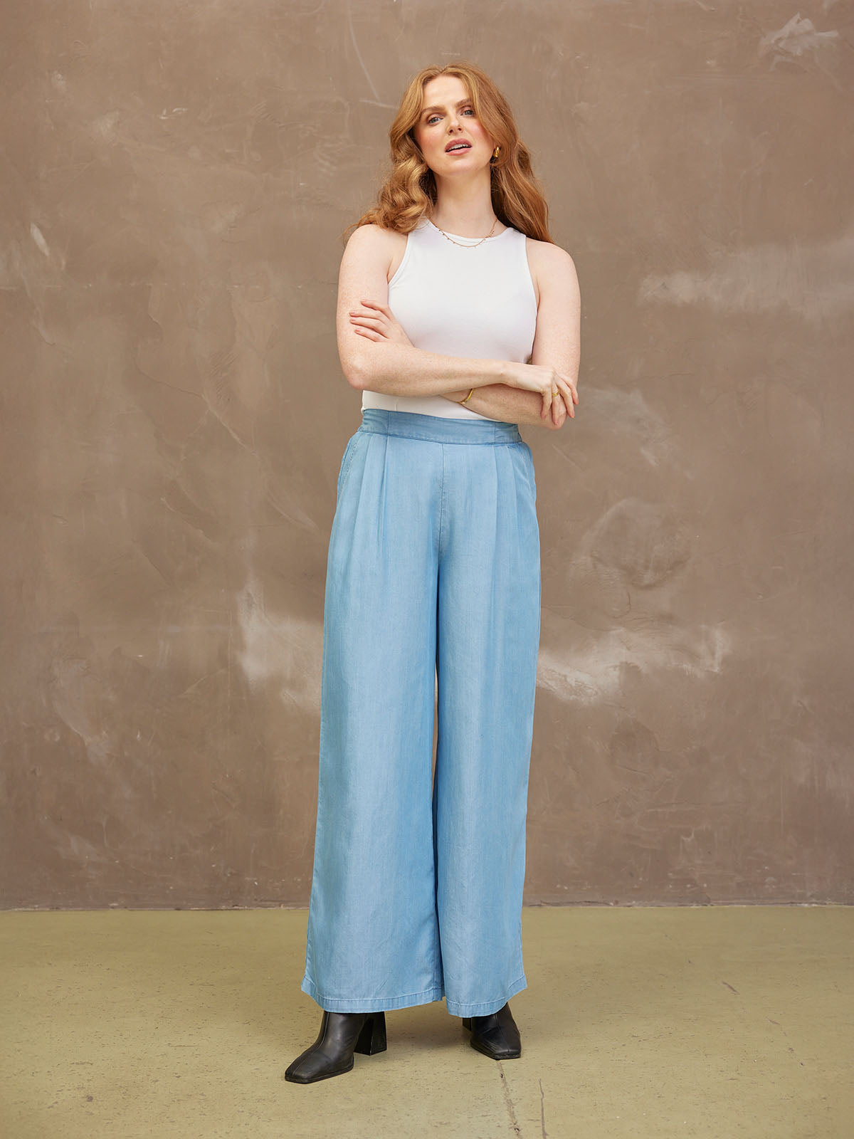 Woman with long hair, wearing a white top and blue wide-leg pants, against a neutral background.