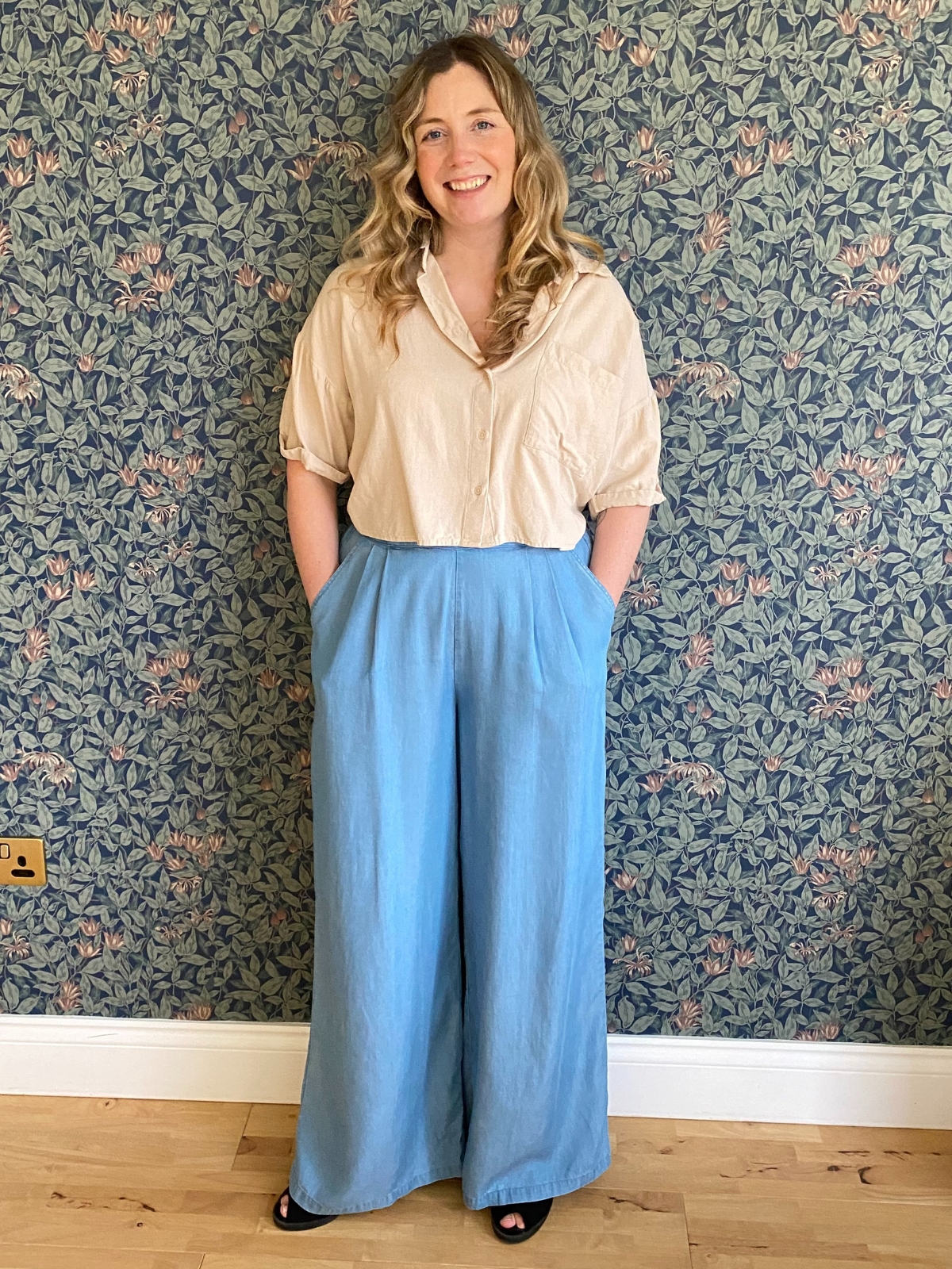 Person in beige shirt and blue pants standing against floral wallpaper.