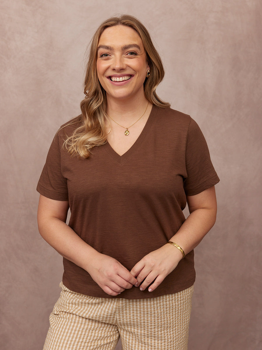 Cathy V-neck Tee in brown, worn by smiling model with striped trousers and gold jewellery, against a dusty pink studio backdrop.