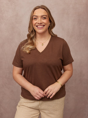 Cathy V-neck Tee in brown, worn by smiling model with striped trousers and gold jewellery, against a dusty pink studio backdrop.