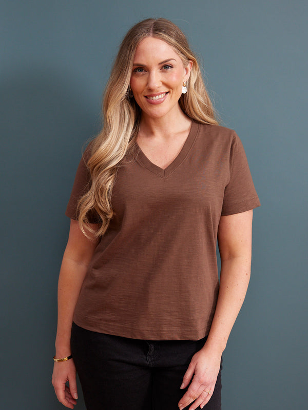 Cathy V-neck Tee in brown, worn by a woman with black jeans against a blue-grey studio background