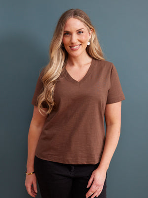 Cathy V-neck Tee in brown, worn by a woman with black jeans against a blue-grey studio background