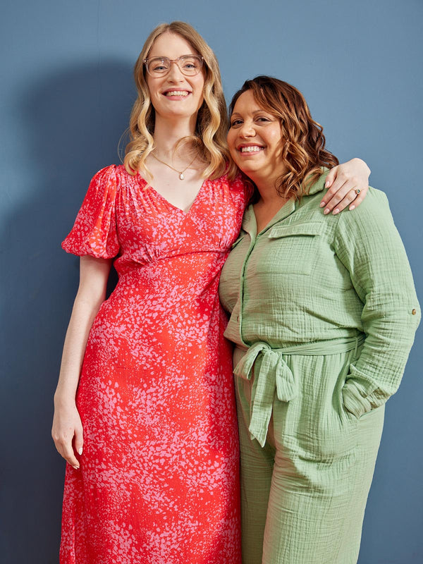 Two women wearing sustainable clothes, including a red and pink sustainable v-neck dress and a green utility jumpsuit, standing in front of a blue wall and smiling at the camera