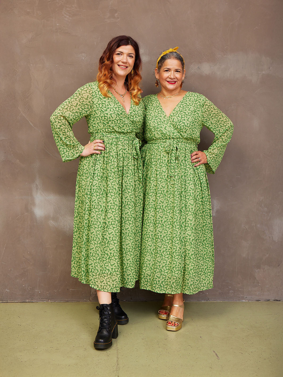 Two people in matching green dresses standing against a textured wall.