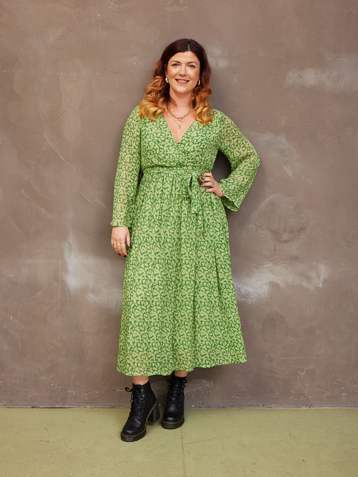 Woman in green patterned dress stands against a textured brown wall.