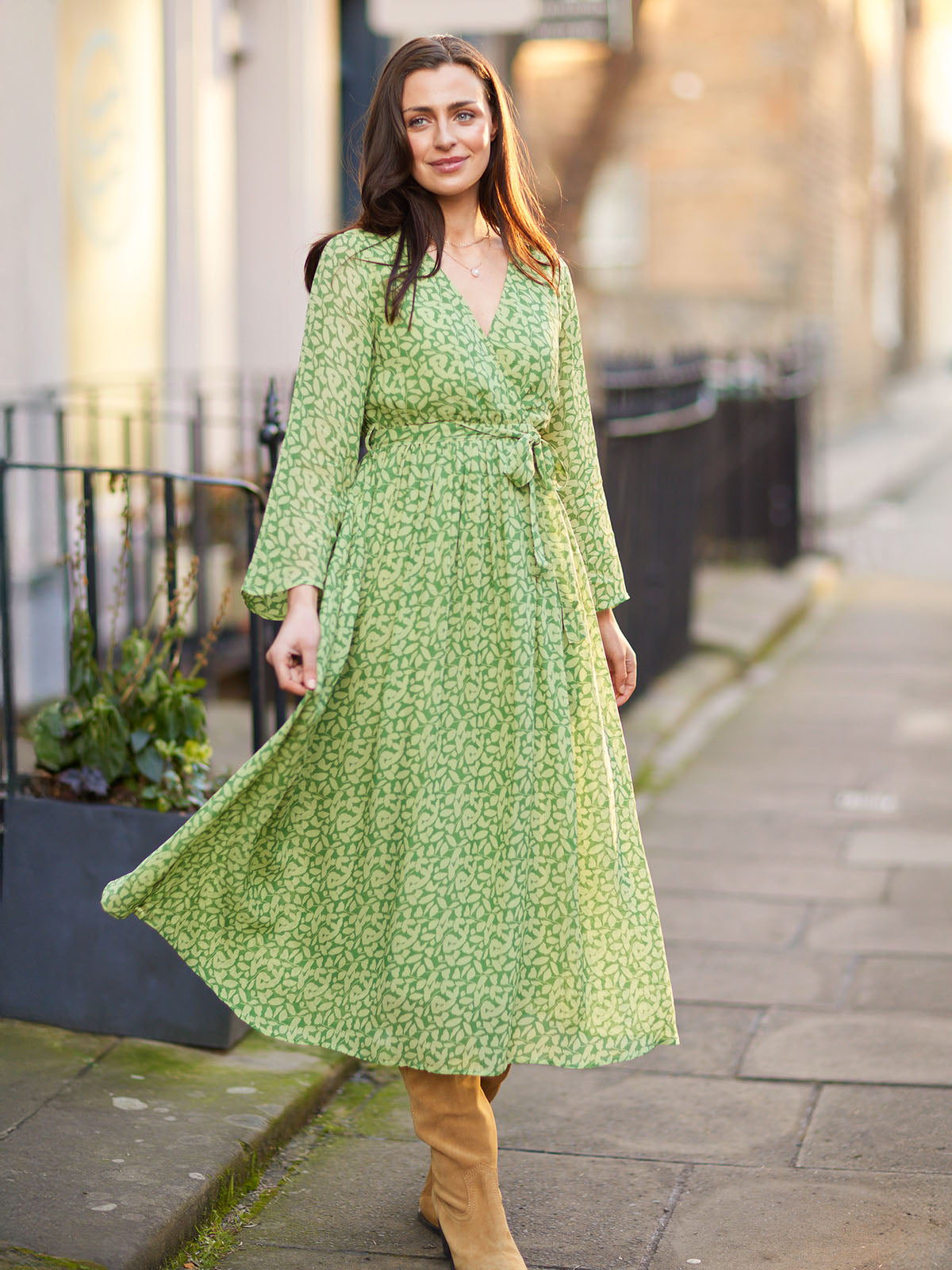 Woman wearing a green patterned dress and boots, walking on a city sidewalk.