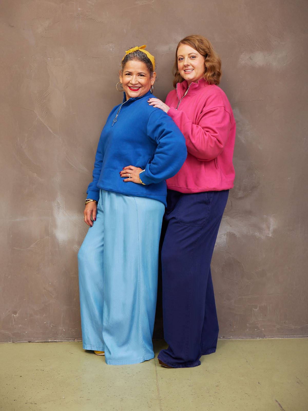 Two smiling women in colorful sweaters and wide-leg pants standing against a neutral background.