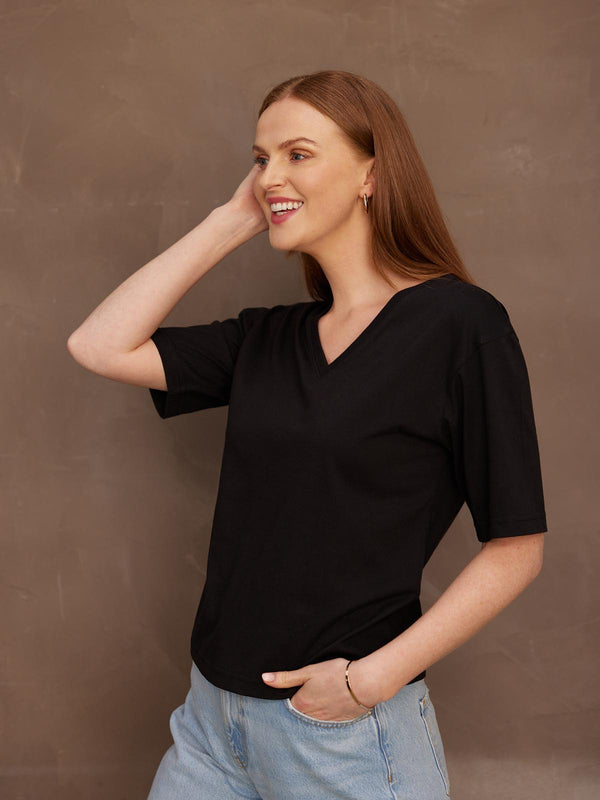 A smiling model looking to the side with her left hand in her pocket and her right hand held up to hear head, wearing the sustainable Freya black t-shirt paired with jeans.