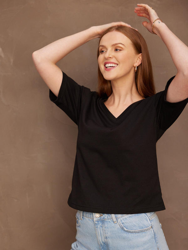 A smiling model looking to the side with both her hands held up to her hair, wearing the sustainable Freya black t-shirt paired with jeans.