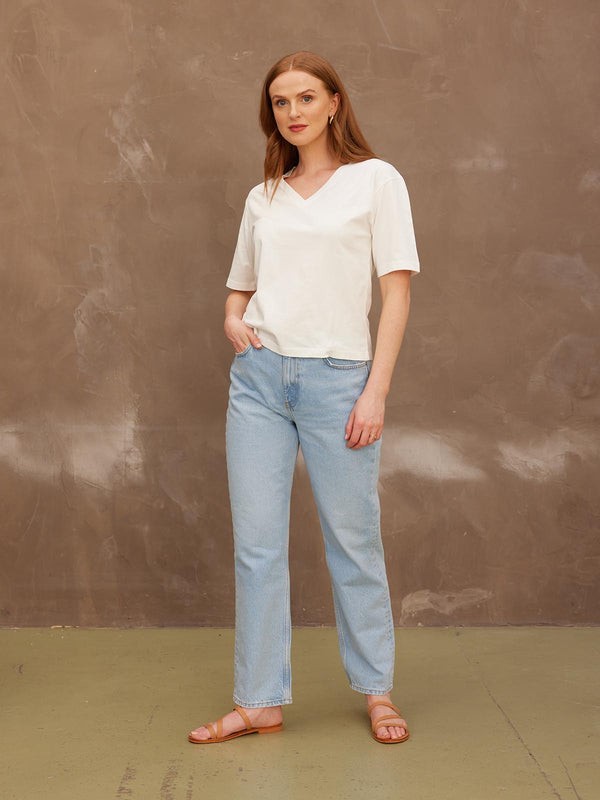 A model looking at the camera with her right hand in her pocket, wearing the sustainable Freya white t-shirt paired with jeans and sandals.