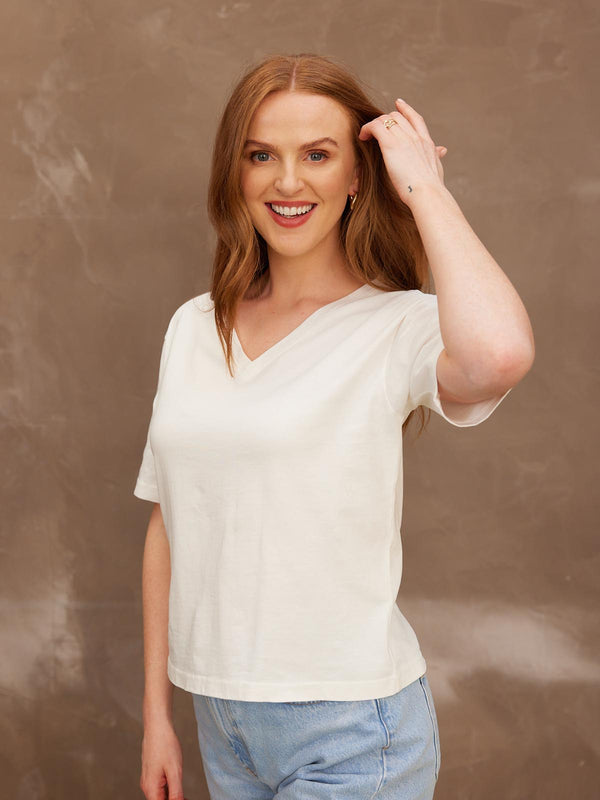 A smiling model looking at the camera with her left hand held up to her hair, wearing the sustainable Freya white t-shirt paired with jeans..