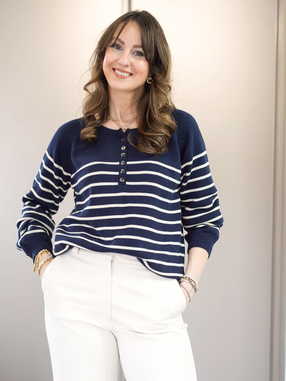 A person in a striped sweater and white pants, smiling with hands in pockets.