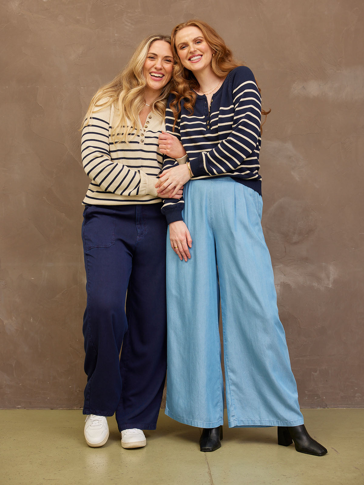 Two people smiling, wearing striped tops and wide-leg trousers, standing together.