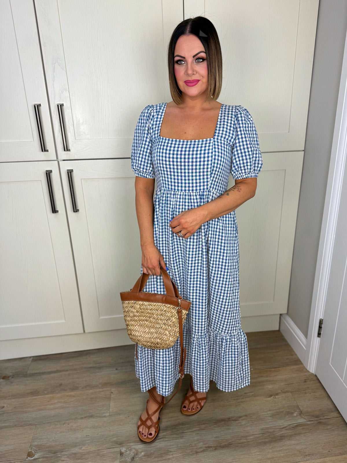 Woman in a blue gingham dress holding a straw handbag, standing indoors.